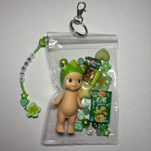 May include: A clear plastic keychain pouch filled with a small doll wearing a green frog hat, miniature snacks, and green beads. A beaded charm with the word "BRAT" and green accents hangs from the pouch.