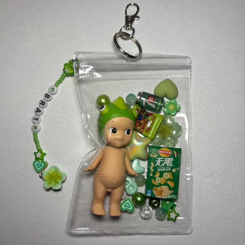 May include: A clear plastic keychain pouch filled with a small doll wearing a green frog hat, miniature snacks, and green beads. A beaded charm with the word "BRAT" and green accents hangs from the pouch.