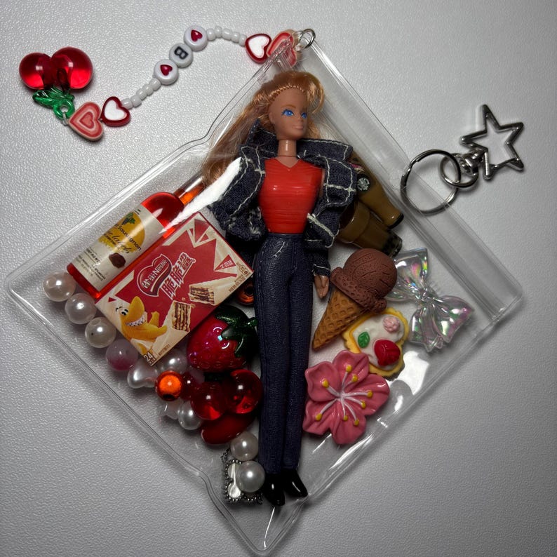 K&ouml;nnte beinhalten: Ein durchsichtiger Kunststoff-Schl&uuml;sselanh&auml;nger mit einer Puppe in rotem Oberteil, dunkler Jeansjacke und Jeans. Der Beutel ist mit Miniaturartikeln gef&uuml;llt, darunter eine Flasche, eine Snackschachtel mit chinesischem Text, eine Erdbeere, ein Eis und eine Blume.