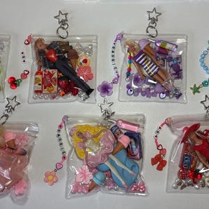 K&ouml;nnte beinhalten: Sieben durchsichtige Kunststoffbeutel, die jeweils eine Miniaturpuppe, verschiedene bunte Perlen und kleine thematische Accessoires enthalten. Jeder Beutel hat einen silbernen sternf&ouml;rmigen Schl&uuml;sselanh&auml;nger und ein perlenbesetztes Anh&auml;ngerband.