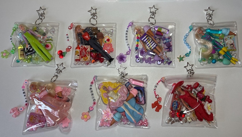 K&ouml;nnte beinhalten: Sieben durchsichtige Kunststoffbeutel, die jeweils eine Miniaturpuppe, verschiedene bunte Perlen und kleine thematische Accessoires enthalten. Jeder Beutel hat einen silbernen sternf&ouml;rmigen Schl&uuml;sselanh&auml;nger und ein perlenbesetztes Anh&auml;ngerband.