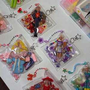 K&ouml;nnte beinhalten: Transparente Kunststoffbeutel mit Miniatur-Modepuppen, bunten Perlen und kleinen Spielzeug-Accessoires. Jeder Beutel verf&uuml;gt &uuml;ber einen sternf&ouml;rmigen Metall-Schl&uuml;sselanh&auml;nger und ein Perlenband mit dem Schriftzug Barbie aus wei&szlig;en Buchstaben.