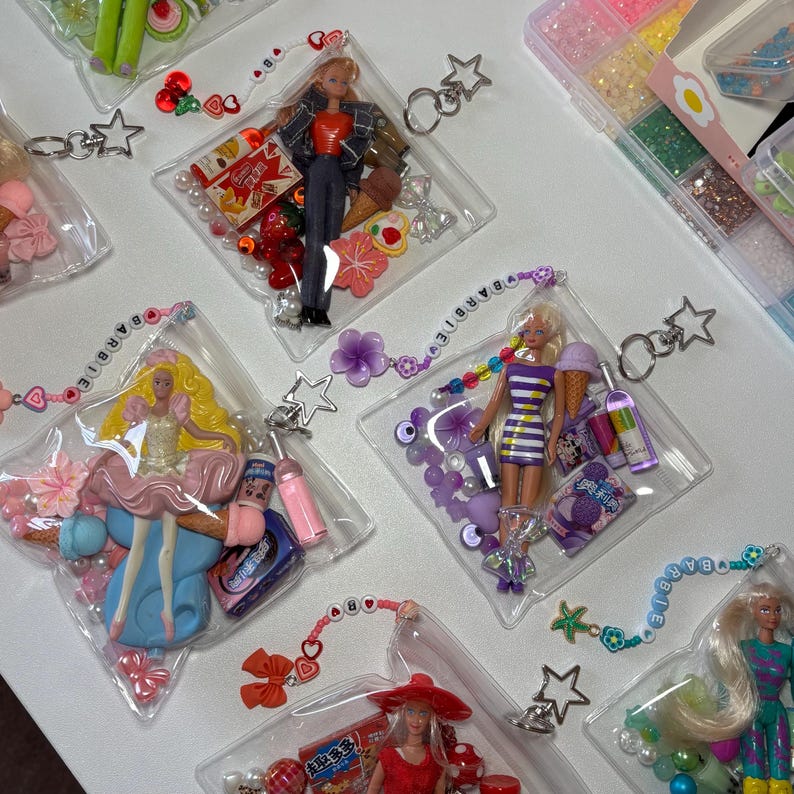 K&ouml;nnte beinhalten: Transparente Kunststoffbeutel mit Miniatur-Modepuppen, bunten Perlen und kleinen Spielzeug-Accessoires. Jeder Beutel verf&uuml;gt &uuml;ber einen sternf&ouml;rmigen Metall-Schl&uuml;sselanh&auml;nger und ein Perlenband mit dem Schriftzug Barbie aus wei&szlig;en Buchstaben.