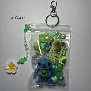 May include: A clear plastic pouch filled with green-themed charms and accessories. Includes a blue Stitch figurine, green beads, a small bottle, and a charm bracelet with a white flower. The pouch is attached to a silver keychain.