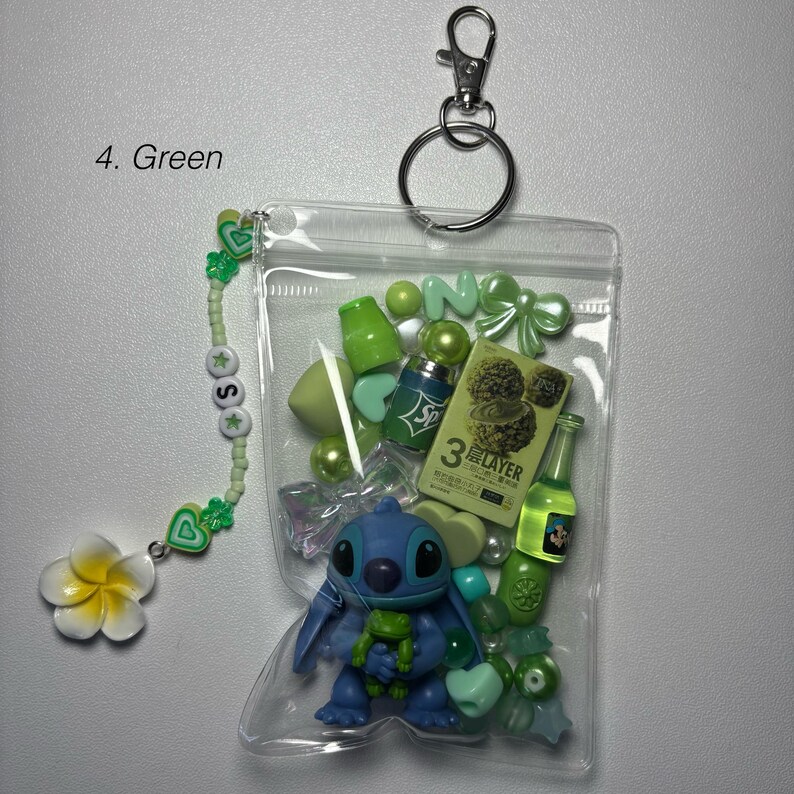 May include: A clear plastic pouch filled with green-themed charms and accessories. Includes a blue Stitch figurine, green beads, a small bottle, and a charm bracelet with a white flower. The pouch is attached to a silver keychain.