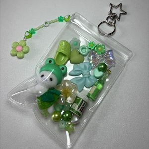 May include: A clear, zippered pouch filled with green and pastel-colored charms and accessories. Includes a frog-themed plushie, beads, bows, flowers, a miniature bottle, and a star-shaped keychain. A beaded charm with a flower is attached.