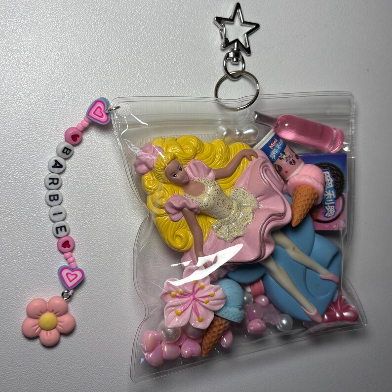 K&ouml;nnte beinhalten: Ein Schl&uuml;sselanh&auml;nger aus durchsichtigem Kunststoffbeutel mit einer blonden Miniaturpuppe in rosa Kleid, einer rosa Flasche, einer Eist&uuml;te und einer kleinen Schachtel. Ein Anh&auml;nger besteht aus rosa Herzperlen, wei&szlig;en Buchstabenperlen BARBIE und einer Blume.