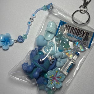 May include: A clear plastic pouch filled with blue charms, including a Stitch figurine, hearts, bows, and a miniature Hershey's bar. A beaded charm with the letter "S" and a flower charm are attached to the pouch, along with a silver keychain.