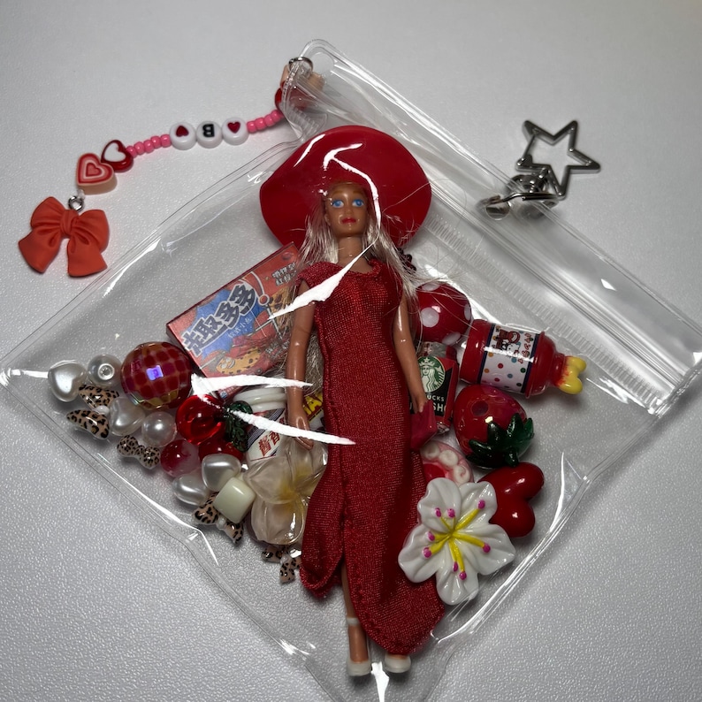 K&ouml;nnte beinhalten: Ein durchsichtiger Kunststoffbeutel mit einer Puppe in rotem Kleid und Hut, umgeben von verschiedenen Anh&auml;ngern, darunter eine rote Schleife, ein Stern, Perlen, eine Erdbeere, eine Blume und eine kleine Schachtel mit japanischem Text.