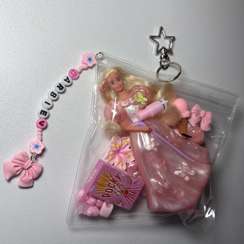 K&ouml;nnte beinhalten: Ein durchsichtiger Kunststoffbeutel mit einer blonden Puppe in einem rosa Kleid, einer Pocky-Miniaturschachtel und kleinen rosa Accessoires. Ein Perlen-Schl&uuml;sselanh&auml;nger mit dem Wort BARBIE, rosa Blumenperlen und einer Schleife ist befestigt.