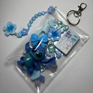 May include: A clear plastic pouch filled with blue-themed charms and accessories. Includes a Stitch figurine, flower and bow-shaped embellishments, a small perfume bottle, and a keychain. The word "STITCH" is spelled out on a beaded chain.