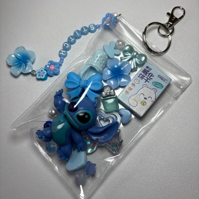 May include: A clear plastic pouch filled with blue-themed charms and accessories. Includes a Stitch figurine, flower and bow-shaped embellishments, a small perfume bottle, and a keychain. The word "STITCH" is spelled out on a beaded chain.