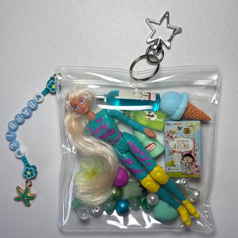 K&ouml;nnte beinhalten: Ein durchsichtiger Kunststoffbeutel mit Rei&szlig;verschluss enth&auml;lt eine blonde Puppe in t&uuml;rkisfarbenem und violettem Outfit, eine blaue Eiswaffel, eine kleine Flasche mit blauer Fl&uuml;ssigkeit und eine Schachtel mit der Aufschrift pejoy. Ein Perlenanh&auml;nger.