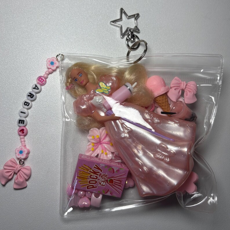 K&ouml;nnte beinhalten: Ein durchsichtiger Kunststoffbeutel-Schl&uuml;sselanh&auml;nger mit einer blonden Puppe in einem gl&auml;nzenden rosa Kleid, einer Pocky-Miniaturpackung, einem rosa Eis, einer rosa Schleife und einer Blumenperle. Eine Perlenkette buchstabiert BARBIE mit einer Schleife.
