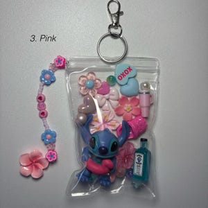 May include: A clear plastic keychain pouch filled with pink and blue charms, including a Stitch figurine, flowers, bows, and a small bottle. The keychain has a beaded charm with pink and blue flowers and the text "XOXO". The text "3. Pink" is visible.