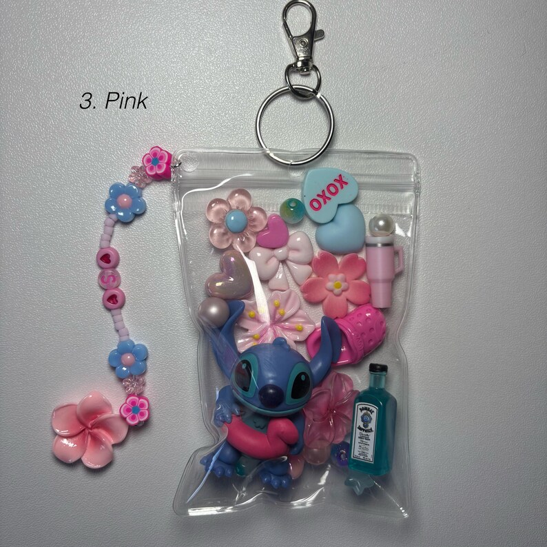 May include: A clear plastic keychain pouch filled with pink and blue charms, including a Stitch figurine, flowers, bows, and a small bottle. The keychain has a beaded charm with pink and blue flowers and the text "XOXO". The text "3. Pink" is visible.
