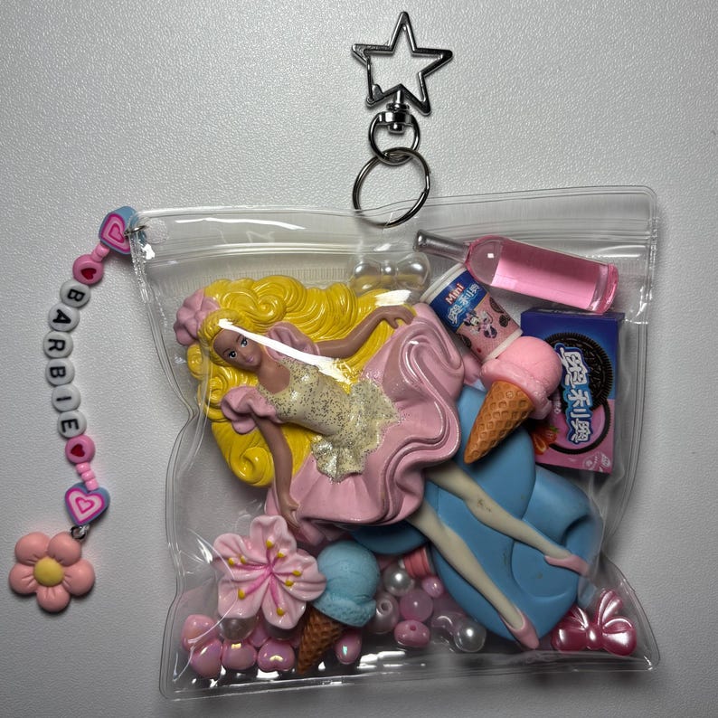 K&ouml;nnte beinhalten: Ein durchsichtiger Kunststoffbeutel-Schl&uuml;sselanh&auml;nger mit sternf&ouml;rmigem Verschluss und einer Perlenkette, die BARBIE buchstabiert, erg&auml;nzt durch Herz- und Blumenanh&auml;nger. Darin befinden sich Miniaturartikel wie eine blonde Puppe und eine Eiswaffel.