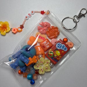 May include: A clear plastic pouch filled with colorful charms, beads, and a keychain. The charms include a blue cartoon character, flowers, hearts, and small food-shaped items. A beaded strand spells out the letter "S".