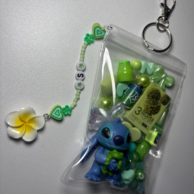 May include: A clear plastic pouch filled with green and blue trinkets, including a Stitch figurine holding a frog. A beaded charm with the letter "S" and a white and yellow flower dangle from a silver clasp.