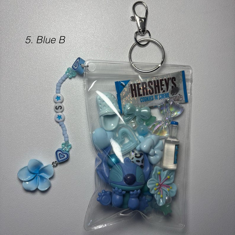 May include: A clear plastic pouch filled with blue charms, including a Stitch figure, bows, and a Hershey's Cookies 'N' Creme candy wrapper. A beaded charm with the letter "S" and a blue flower dangle from the pouch.