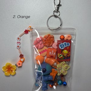 May include: A clear, zippered pouch filled with colorful charms and a keychain. Charms include a blue Stitch figurine, orange and pink sweets, and a red snack box. A beaded charm with the letter "S" and a yellow flower dangle from the pouch.