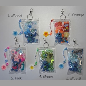 May include: Five clear zipper pouches with keychains, each filled with colorful beads, charms, and a Stitch figurine. The pouches are labeled: Blue A, Orange, Pink, Green, and Blue B. Each pouch has a beaded charm strap.