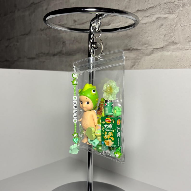 May include: A clear plastic bag keychain filled with a small doll wearing a green frog hat, miniature soda cans, and other green accessories. The keychain is suspended from a silver metal stand.