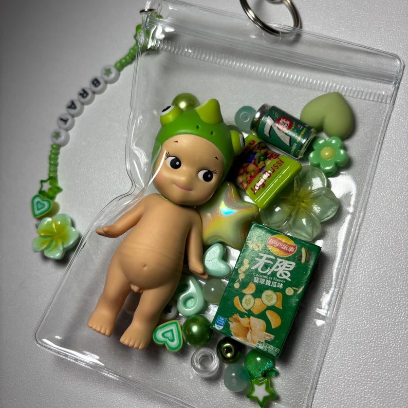 May include: A clear plastic pouch filled with a small doll wearing a green frog hat, miniature snacks, and green beads. A beaded bracelet spelling "BRAT" is attached to the pouch. The pouch is secured with a silver keyring.