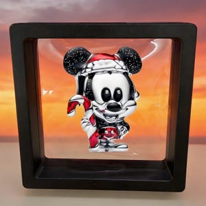 May include: A silver Mickey Mouse figurine in a black display case. Mickey is wearing a Santa hat and holding a red and white candy cane. The background is a gradient of orange and yellow.