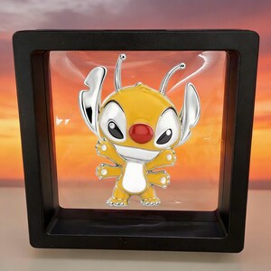 May include: A yellow and silver enamel pin featuring a four-armed creature with large ears, antennae, and a red nose, suspended inside a square black floating display frame with a clear plastic membrane holding the pin in the center.