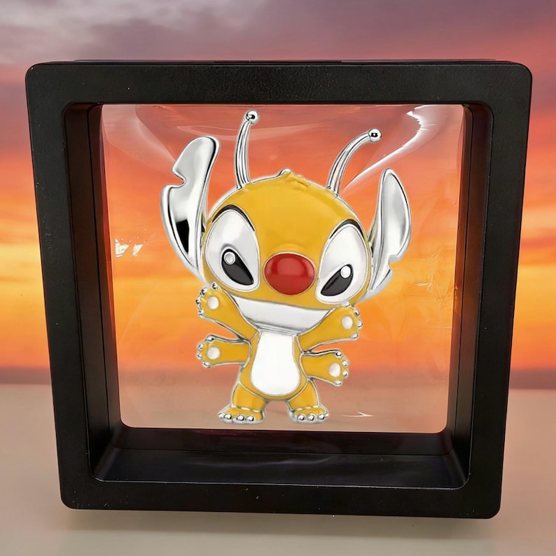 May include: A yellow and silver enamel pin featuring a four-armed creature with large ears, antennae, and a red nose, suspended inside a square black floating display frame with a clear plastic membrane holding the pin in the center.