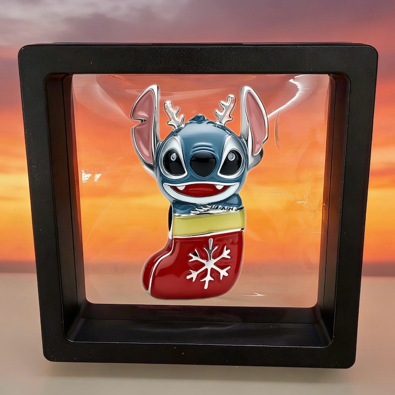 May include: A blue enamel charm featuring the character Stitch with silver reindeer antlers, emerging from a red Christmas stocking decorated with a white snowflake, suspended within a square black floating display frame.