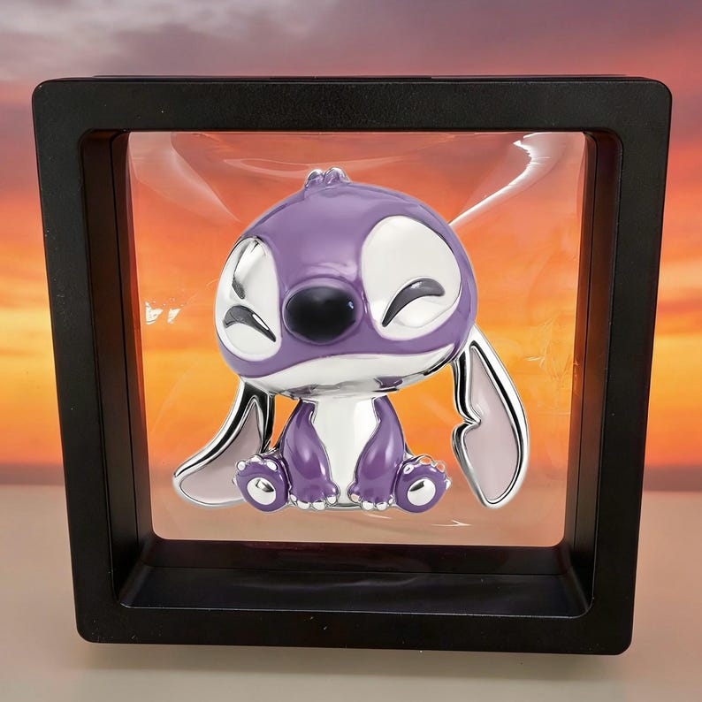 May include: A purple and silver enamel pin featuring a cartoon character with large eyes, a black nose, and long ears, suspended inside a square black floating display frame with a clear plastic membrane holding the pin in place.