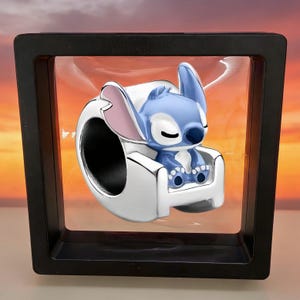 May include: A blue and white charm featuring a sleeping cartoon character in a silver chair. The charm is displayed in a black box with a clear front, set against a sunset background. The character has large ears and is in a relaxed pose.