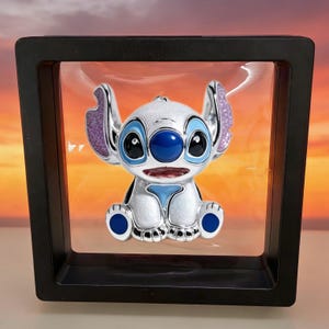 May include: A white and blue enamel pin of the character Stitch, featuring glittery purple inner ears and a glossy finish, suspended inside a square black floating display frame with a clear plastic membrane holding the pin in place.