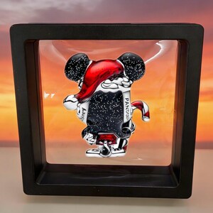 May include: A Mickey Mouse charm in a black display case. The charm is black with white speckles, a red Santa hat, and a red and white candy cane. The charm has the word "Pandora" on it.