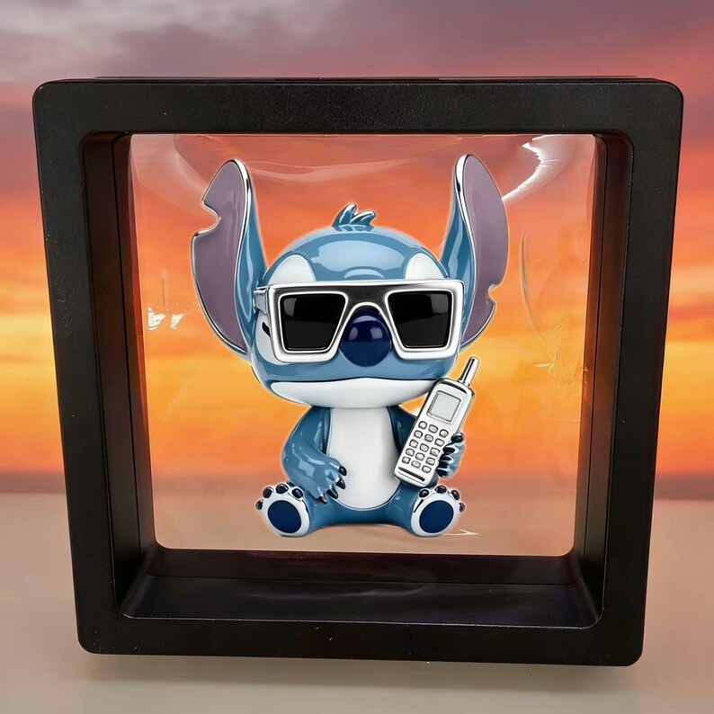 May include: A blue and white alien character figurine with large ears, wearing black sunglasses and holding a small gray mobile phone. The figure is suspended inside a square black frame with a clear plastic display window.