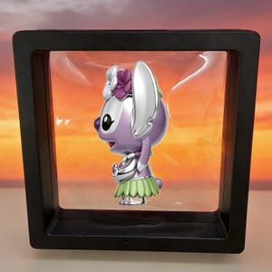 May include: A purple and silver character figurine with large ears, wearing a green grass-style skirt and a purple flower accessory on its head, suspended inside a square black floating display frame with a clear plastic membrane.