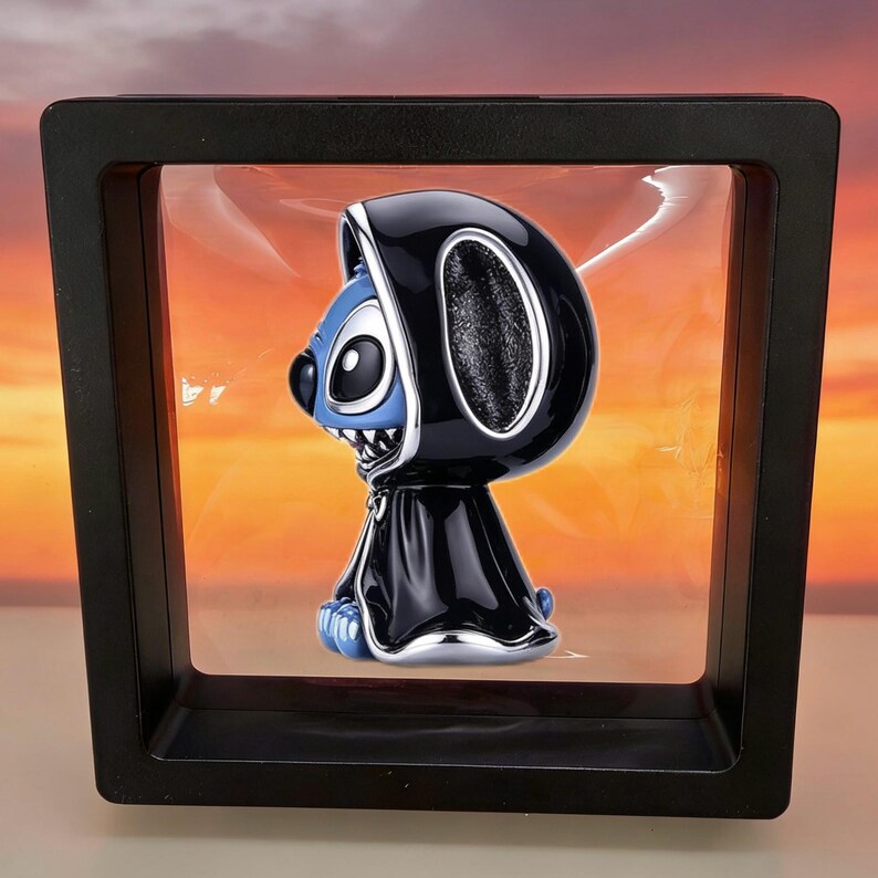 May include: A blue character figurine with large ears and a black nose, wearing a black hooded cloak with silver trim. The figurine is suspended inside a square black floating display frame with a clear membrane, set against a sunset background.