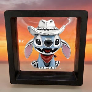 May include: A silver-toned charm featuring a light blue character with large black eyes, a wide grin, and pink inner ears. The character wears a silver cowboy hat and a red bandana around its neck, suspended within a square black floating display frame.