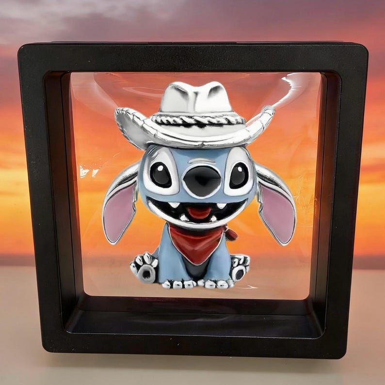 May include: A silver-toned charm featuring a light blue character with large black eyes, a wide grin, and pink inner ears. The character wears a silver cowboy hat and a red bandana around its neck, suspended within a square black floating display frame.