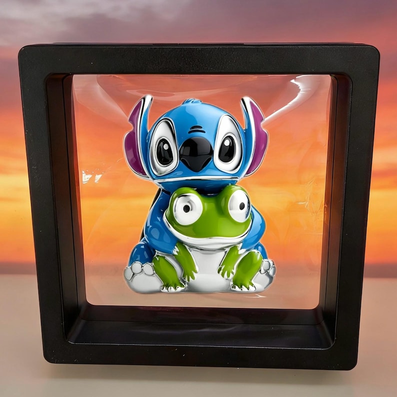 May include: A glossy enamel pin featuring a blue alien character with large ears hugging a small green frog. The pin is suspended within a square black floating display frame, showcasing a vibrant, smooth finish and detailed character features.