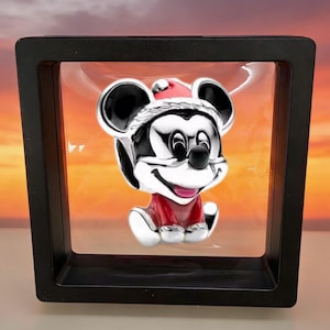 May include: A silver-toned charm featuring a character with large round ears, wearing a red and white Santa hat and a red outfit, suspended within a square black floating display frame with a clear membrane holding the piece in the center.