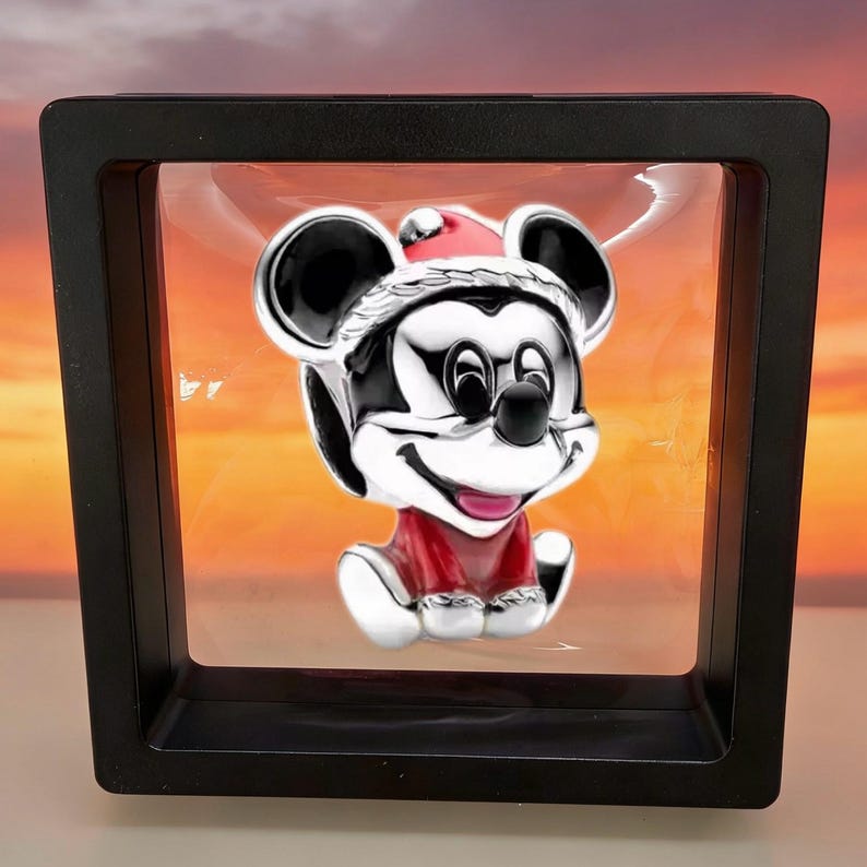 May include: A silver-toned charm featuring a character with large round ears, wearing a red and white Santa hat and a red outfit, suspended within a square black floating display frame with a clear membrane holding the piece in the center.