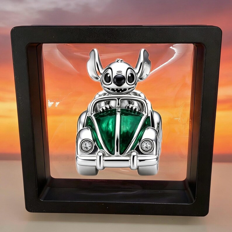 Op de afbeelding: Een zilverkleurige bedel met het personage Stitch in een groene auto met heldere kristallen koplampen. De bedel hangt in een vierkant zwart zwevend displayframe en toont een gedetailleerde metalen afwerking en levendig groen emaille.