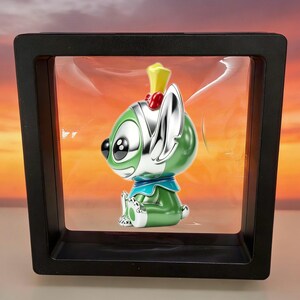 May include: A green and silver cartoon character figurine with large black eyes, a yellow crown, and a blue collar, suspended inside a square black floating display frame with a clear plastic membrane that holds the object in place.