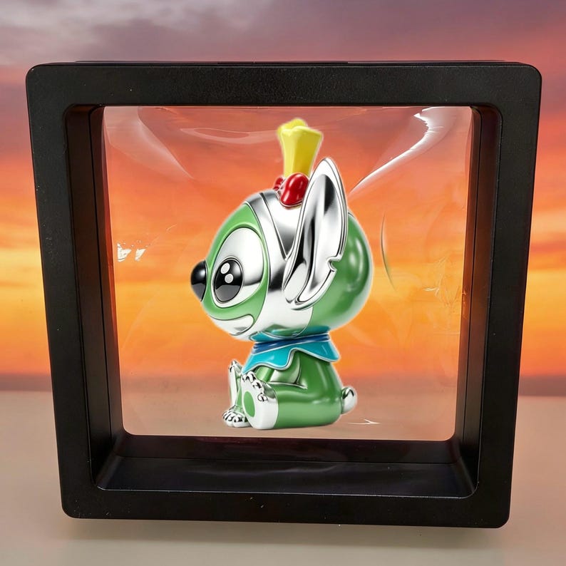 May include: A green and silver cartoon character figurine with large black eyes, a yellow crown, and a blue collar, suspended inside a square black floating display frame with a clear plastic membrane that holds the object in place.