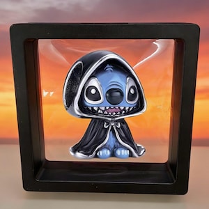 May include: A blue character with large black eyes and a wide grin wearing a black hooded cloak, suspended inside a square black floating display frame. The character features pointed ears and a small bow detail at the neck of the dark cape.