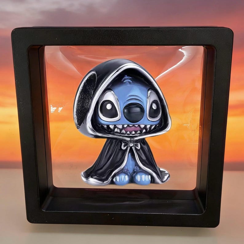 May include: A blue character with large black eyes and a wide grin wearing a black hooded cloak, suspended inside a square black floating display frame. The character features pointed ears and a small bow detail at the neck of the dark cape.
