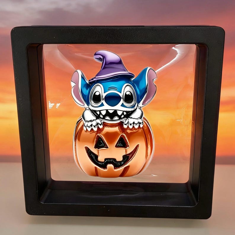 May include: A blue character with large ears and a purple witch hat sits atop a metallic orange jack-o'-lantern. The figure is encased within a square black floating display frame, creating a suspended effect against a sunset-colored background.